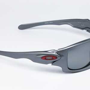 May include: Gray sunglasses with a wraparound design. The frames are a matte gray with a red logo on the side. The lenses are a dark gray color, and the arms curve for a secure fit. The sunglasses are set against a white background.