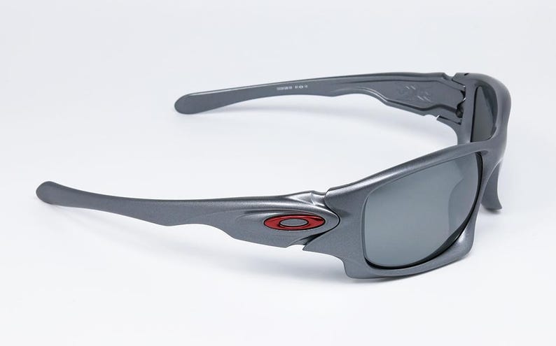 May include: Gray sunglasses with a wraparound design. The frames are a matte gray with a red logo on the side. The lenses are a dark gray color, and the arms curve for a secure fit. The sunglasses are set against a white background.