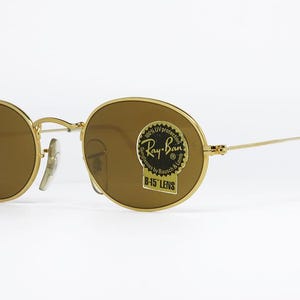 Pu&ograve; includere: Occhiali da sole con montatura dorata e lenti marroni. Le lenti ovali sono inserite in una montatura in metallo dorato. Gli occhiali da sole hanno un adesivo Ray-Ban sulla lente. Su sfondo bianco.