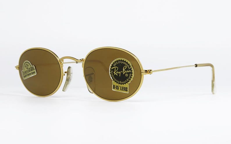 Pu&ograve; includere: Occhiali da sole con montatura dorata e lenti marroni. Le lenti ovali sono inserite in una montatura in metallo dorato. Gli occhiali da sole hanno un adesivo Ray-Ban sulla lente. Su sfondo bianco.