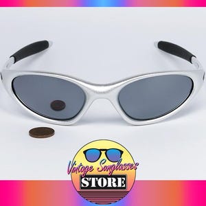 May include: Silver-framed sunglasses with dark gray lenses. The sunglasses have black accents on the temples. A penny and a logo for "Vintage Sunglasses Store" are also in the image.