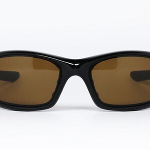 May include: Black sunglasses with brown tinted lenses. The sunglasses have a curved, wraparound design with a thick frame. The lenses are a warm brown colour, and the frame is glossy black.