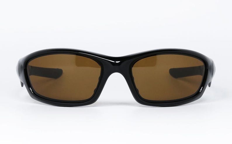 May include: Black sunglasses with brown tinted lenses. The sunglasses have a curved, wraparound design with a thick frame. The lenses are a warm brown colour, and the frame is glossy black.