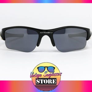 K&ouml;nnte beinhalten: Schwarze Oakley-Sonnenbrille mit grauen Gl&auml;sern. Die Sonnenbrille hat ein schlankes, modernes Design mit einem geschwungenen Rahmen und einem dezenten Oakley-Logo auf der Br&uuml;cke. Das Bild enth&auml;lt ein Logo f&uuml;r Vintage Sunglasses Store.