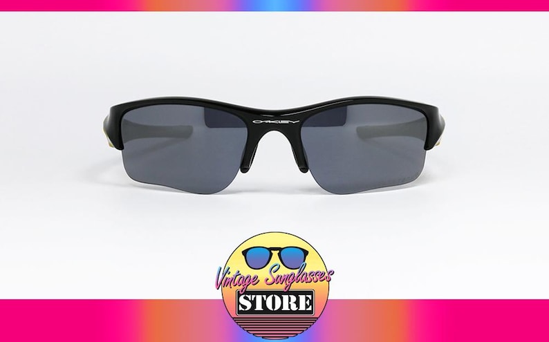 K&ouml;nnte beinhalten: Schwarze Oakley-Sonnenbrille mit grauen Gl&auml;sern. Die Sonnenbrille hat ein schlankes, modernes Design mit einem geschwungenen Rahmen und einem dezenten Oakley-Logo auf der Br&uuml;cke. Das Bild enth&auml;lt ein Logo f&uuml;r Vintage Sunglasses Store.