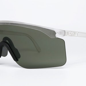 Puede incluir: Gafas de sol con montura blanca y lentes grises oscuras con la palabra "Oakley" impresa en el lateral de la montura.