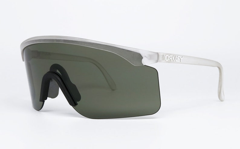 Puede incluir: Gafas de sol con montura blanca y lentes grises oscuras con la palabra "Oakley" impresa en el lateral de la montura.