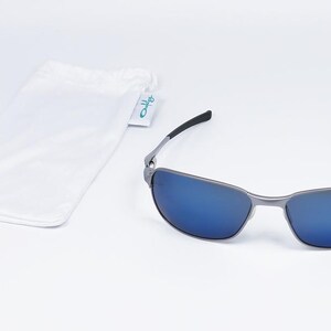 May include: Sunglasses with a silver frame and blue lenses. The sunglasses have black arms and are accompanied by a white drawstring pouch with a small tag. The tag has the word "Oakley" on it.