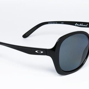 Puede incluir: Gafas de sol negras con montura grande y redondeada y lentes oscuras. Las patillas son elegantes y llevan el nombre de la marca "Backhand". El logotipo de Oakley es visible en las patillas. Un accesorio elegante para la protecci&oacute;n ocular.
