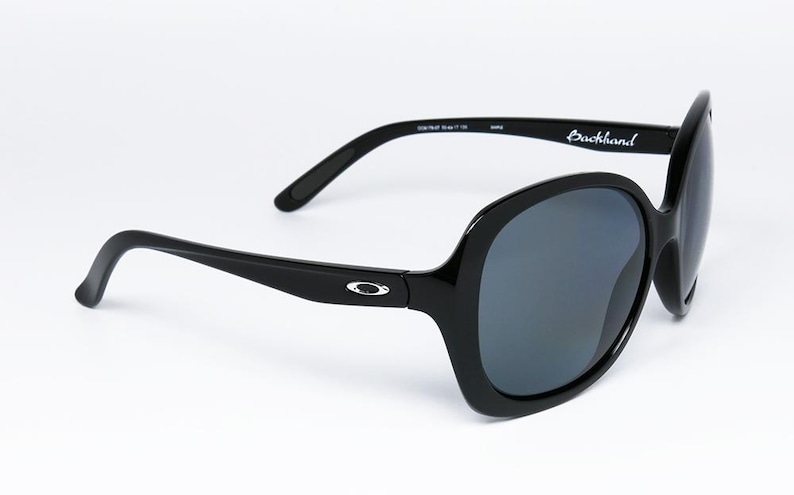 Puede incluir: Gafas de sol negras con montura grande y redondeada y lentes oscuras. Las patillas son elegantes y llevan el nombre de la marca "Backhand". El logotipo de Oakley es visible en las patillas. Un accesorio elegante para la protecci&oacute;n ocular.