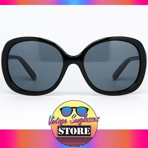 Puede incluir: Gafas de sol negras de gran tama&ntilde;o con lentes de color gris oscuro. Las gafas de sol tienen una montura redondeada en forma de mariposa. La imagen incluye un logotipo que dice "Vintage Sunglasses Store".
