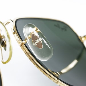May include: Close-up of gold-tone aviator sunglasses with green lenses. The sunglasses have a metal frame and a clear plastic nose bridge. The sunglasses are made in the USA.