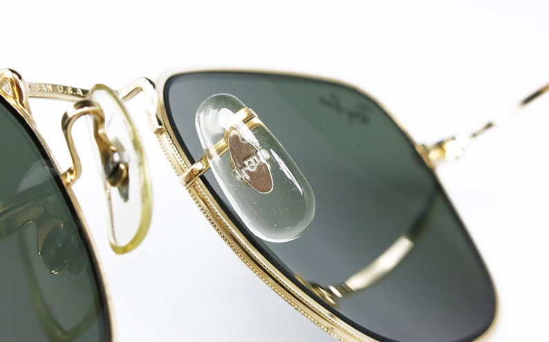 May include: Close-up of gold-tone aviator sunglasses with green lenses. The sunglasses have a metal frame and a clear plastic nose bridge. The sunglasses are made in the USA.
