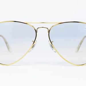 Op de afbeelding: Gouden aviator zonnebril met lichtblauwe gradient lenzen. De zonnebril heeft een Ray-Ban logo op de rechter lens.