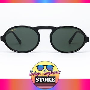 May include: Black Ray-Ban sunglasses with oval lenses and a bridge connecting the frames. The lenses are dark green, and the frames have a glossy finish. The sunglasses are displayed on a white background. The logo of the Vintage Sunglasses Store is at the bottom.