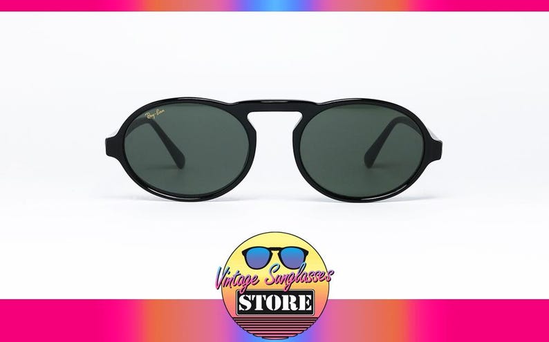 May include: Black Ray-Ban sunglasses with oval lenses and a bridge connecting the frames. The lenses are dark green, and the frames have a glossy finish. The sunglasses are displayed on a white background. The logo of the Vintage Sunglasses Store is at the bottom.