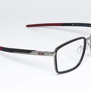 May include: Eyeglasses with a black frame and clear lenses. The temples are black with red tips. The frame has a silver bridge and the Oakley logo on the side. The glasses are set against a white background.