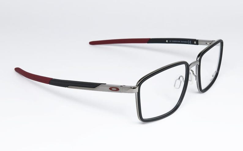 May include: Eyeglasses with a black frame and clear lenses. The temples are black with red tips. The frame has a silver bridge and the Oakley logo on the side. The glasses are set against a white background.