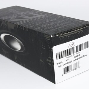 May include: A black rectangular box with a silver oval design and a white label. The label includes a barcode and text that reads "OX3235 3218 Oph. Spindle (54) Gunmetal/Ste Black". The box has a textured pattern.