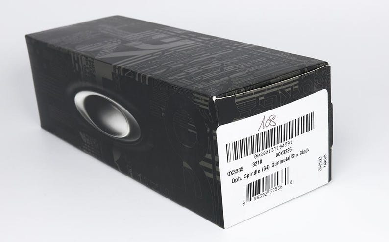 May include: A black rectangular box with a silver oval design and a white label. The label includes a barcode and text that reads "OX3235 3218 Oph. Spindle (54) Gunmetal/Ste Black". The box has a textured pattern.