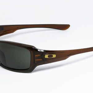 May include: Brown sunglasses with dark lenses. The rectangular frames have a gold logo on the temple. The arms curve back from the frame. The sunglasses are set against a white background.