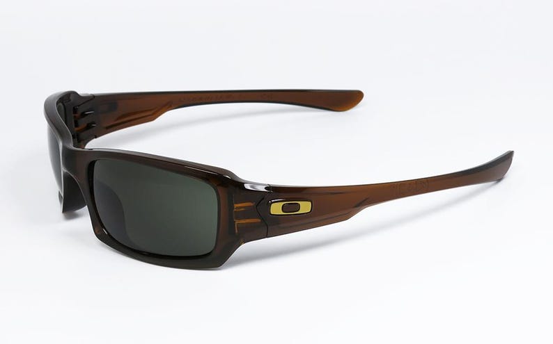 May include: Brown sunglasses with dark lenses. The rectangular frames have a gold logo on the temple. The arms curve back from the frame. The sunglasses are set against a white background.