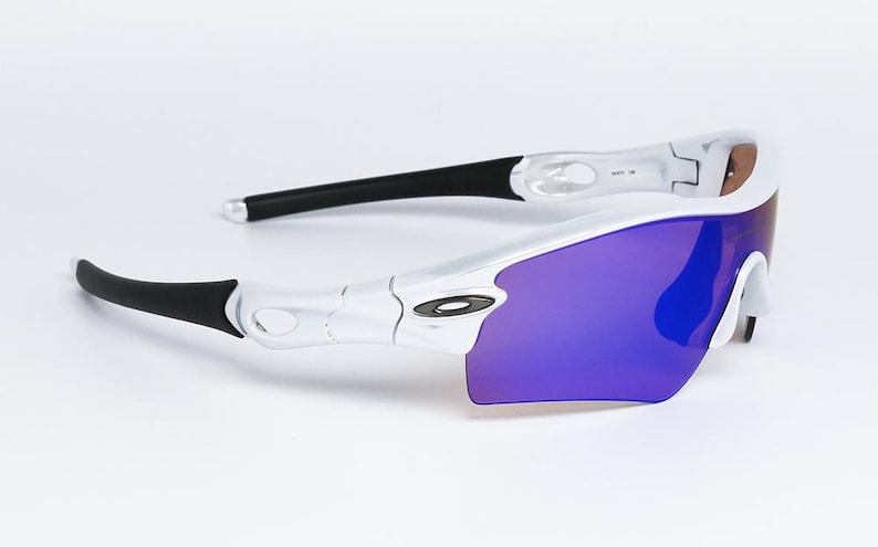 Pu&ograve; includere: Occhiali da sole sportivi bianchi e neri con lenti viola. Gli occhiali hanno un design elegante e moderno con una montatura curva e aste nere. Le lenti sono di un vibrante colore viola.