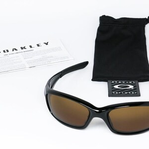 May include: Black Oakley sunglasses with brown lenses, a black drawstring bag, and a warranty card. The sunglasses have a sleek, modern design. The Oakley logo is visible on the card and the bag's tag.