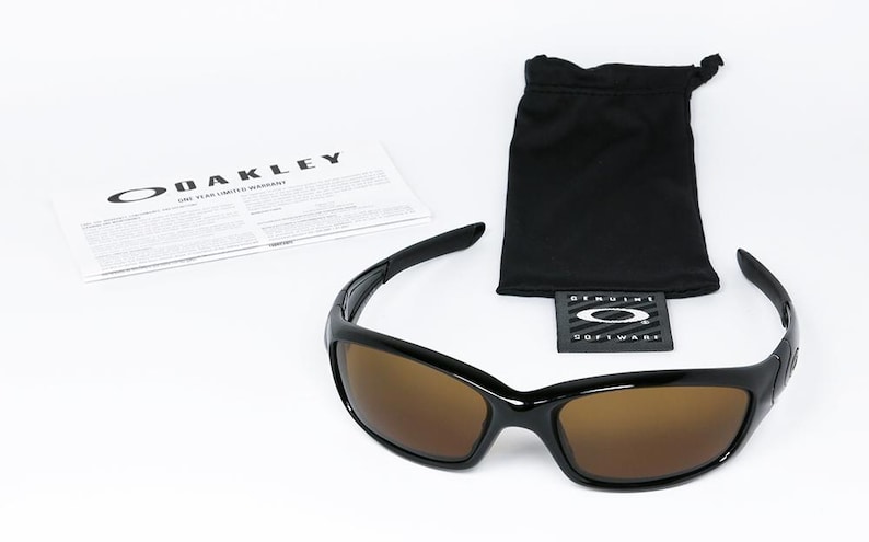 May include: Black Oakley sunglasses with brown lenses, a black drawstring bag, and a warranty card. The sunglasses have a sleek, modern design. The Oakley logo is visible on the card and the bag's tag.