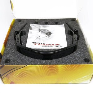 May include: Black Oakley sunglasses in an open box with a foam insert. The sunglasses have a sleek, modern design. A white pamphlet with the Oakley logo is visible. The box has a gold and brown design.