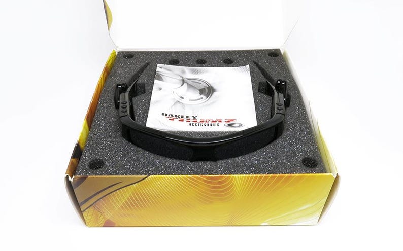 May include: Black Oakley sunglasses in an open box with a foam insert. The sunglasses have a sleek, modern design. A white pamphlet with the Oakley logo is visible. The box has a gold and brown design.
