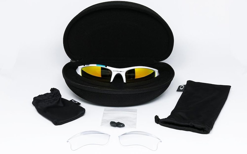 May include: White-framed sunglasses with yellow mirrored lenses rest in an open black case. The image includes a black drawstring bag, a microfiber pouch, a small bag with nose pads, and two clear replacement lenses. The sunglasses are a sporty design.