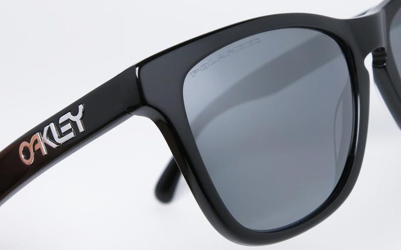 May include: Close-up of black Oakley sunglasses with gray polarized lenses. The Oakley logo is visible on the temple, and the word "POLARIZED" is printed on the lens. The sunglasses have a square frame.