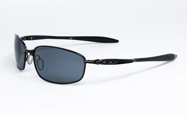 Puede incluir: Gafas de sol negras con un dise&ntilde;o elegante y moderno. Las lentes son de color gris oscuro y las monturas de metal negro. Las patillas se curvan suavemente y las almohadillas nasales son ajustables. Sobre fondo blanco.