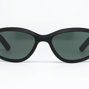 Pode incluir: &Oacute;culos de sol pretos com acabamento fosco. As arma&ccedil;&otilde;es ovais t&ecirc;m lentes verde-escuras e o logotipo da Ray-Ban na haste esquerda. Os &oacute;culos est&atilde;o em um fundo branco.