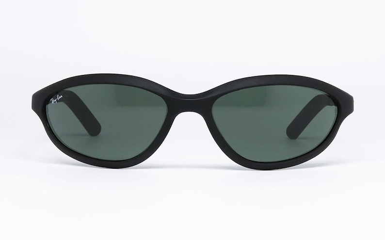 Pode incluir: &Oacute;culos de sol pretos com acabamento fosco. As arma&ccedil;&otilde;es ovais t&ecirc;m lentes verde-escuras e o logotipo da Ray-Ban na haste esquerda. Os &oacute;culos est&atilde;o em um fundo branco.