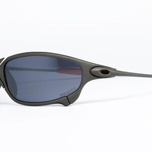 May include: Dark gray sunglasses with a curved frame and dark lenses. The temples are long and gray, with red tips. The sunglasses have a sporty, modern design, suitable for outdoor activities.