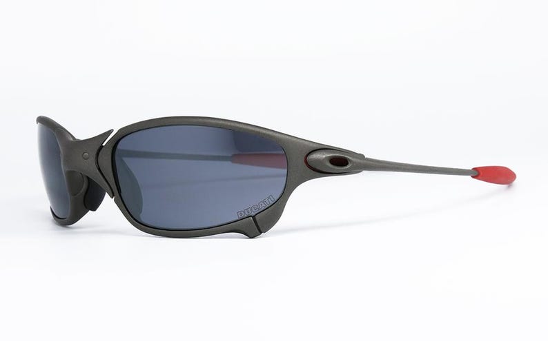 May include: Dark gray sunglasses with a curved frame and dark lenses. The temples are long and gray, with red tips. The sunglasses have a sporty, modern design, suitable for outdoor activities.