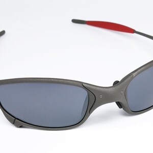 May include: Gray sunglasses with dark lenses and red accents on the temples. The sporty eyewear features a wraparound design and a matte finish, suitable for outdoor activities and everyday wear.
