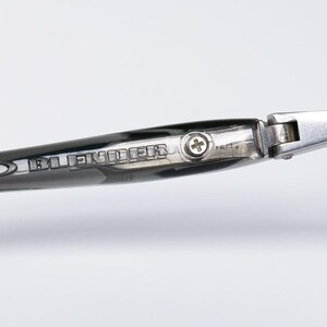 May include: Close-up of sunglasses with a dark frame and lenses. The temple arm is black with the word "BLENDER" printed in silver. A silver hinge connects the temple to the frame. The sunglasses have a modern, sleek design.