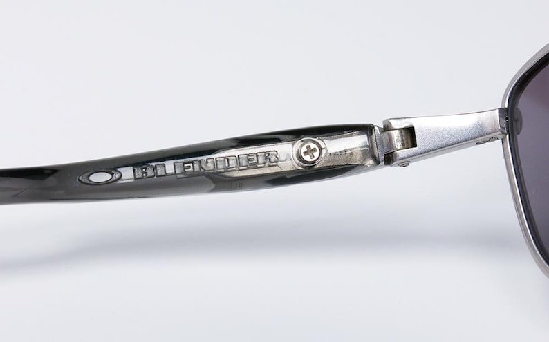 May include: Close-up of sunglasses with a dark frame and lenses. The temple arm is black with the word "BLENDER" printed in silver. A silver hinge connects the temple to the frame. The sunglasses have a modern, sleek design.