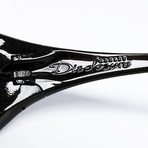 K&ouml;nnte beinhalten: Nahaufnahme einer schwarzen Sonnenbrille mit braunen Gl&auml;sern. Der Rahmen hat ein gl&auml;nzendes Finish und der Text "Oakley Disclosure" ist auf dem B&uuml;gel sichtbar. Die Sonnenbrille ist f&uuml;r Outdoor-Aktivit&auml;ten konzipiert.