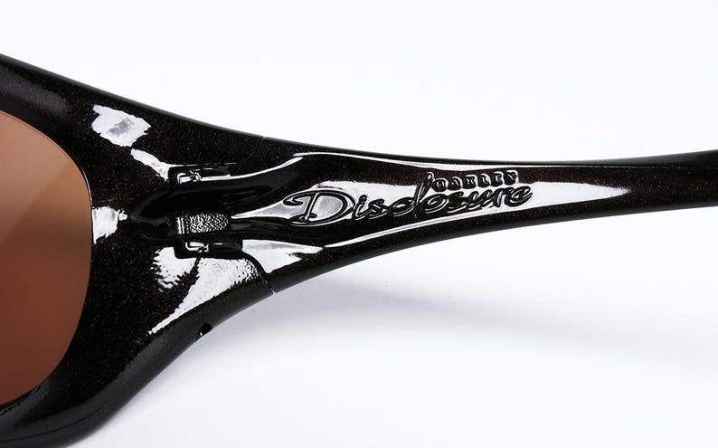 K&ouml;nnte beinhalten: Nahaufnahme einer schwarzen Sonnenbrille mit braunen Gl&auml;sern. Der Rahmen hat ein gl&auml;nzendes Finish und der Text "Oakley Disclosure" ist auf dem B&uuml;gel sichtbar. Die Sonnenbrille ist f&uuml;r Outdoor-Aktivit&auml;ten konzipiert.