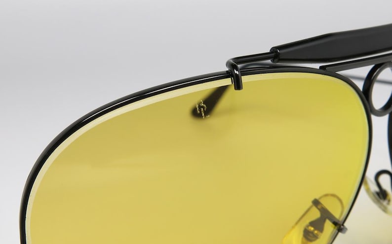 Puede incluir: Primer plano de gafas de sol estilo aviador con lentes amarillas y montura de metal negro. Las lentes tienen una ligera curvatura y la montura presenta un puente y patillas. Dise&ntilde;o cl&aacute;sico.