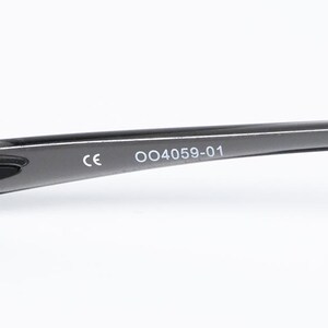 May include: Close-up of a black eyeglass frame arm. The arm has the text "CE" and "004059-01" printed in white. The frame is curved and appears to be made of a smooth, glossy material.