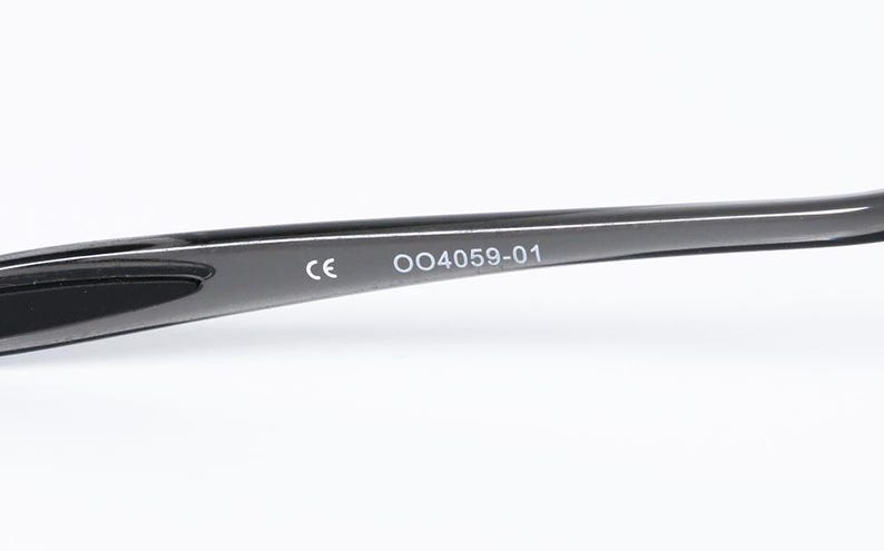 May include: Close-up of a black eyeglass frame arm. The arm has the text "CE" and "004059-01" printed in white. The frame is curved and appears to be made of a smooth, glossy material.