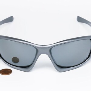May include: Gray sunglasses with a curved frame and gray lenses. A penny is next to the sunglasses. The sunglasses have a sporty design and are suitable for outdoor activities.