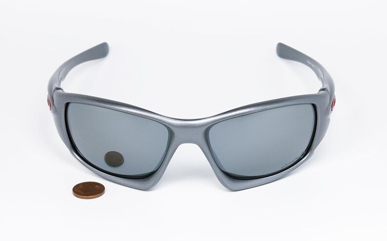 May include: Gray sunglasses with a curved frame and gray lenses. A penny is next to the sunglasses. The sunglasses have a sporty design and are suitable for outdoor activities.