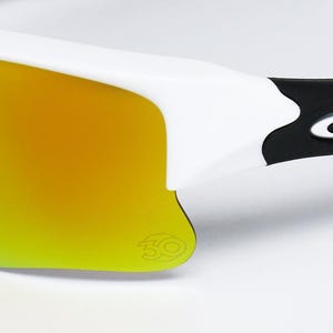 May include: Close-up of white sunglasses with a yellow-orange gradient lens. The frame is white with a black accent near the temple, featuring a brand logo. The lens has a subtle logo in the corner.