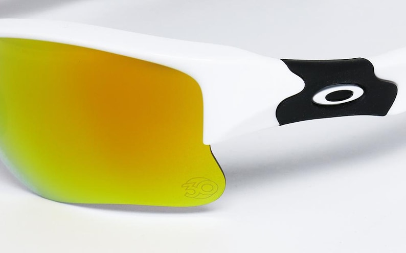 May include: Close-up of white sunglasses with a yellow-orange gradient lens. The frame is white with a black accent near the temple, featuring a brand logo. The lens has a subtle logo in the corner.